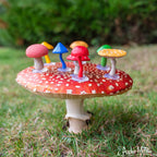 A collection of mini mushrooms in vibrant colors decorates a large red mushroom cap, surrounded by grass.