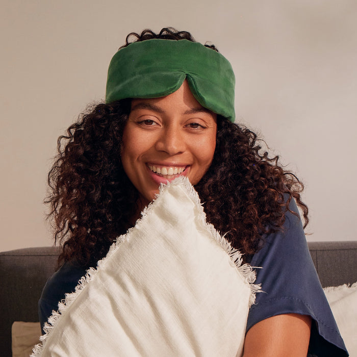 A smiling person with curly hair wears the Arey x Lunya Silk Sleep Mask by Arey in green on their forehead and hugs a white fringed pillow while sitting on a couch.