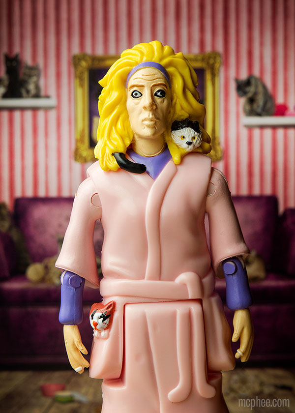 The Crazy Cat Lady® Action Figure captures the quirky essence of a woman with messy hair in a pink robe, surrounded by six cats in a room featuring striped walls and a purple sofa.