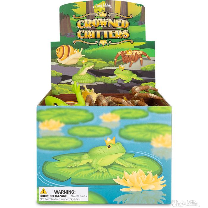 A box of vinyl toys called "Crowned Critters" showcases forest critters, featuring a frog donning a crown on lily pads. It comes with a warning label indicating small parts.