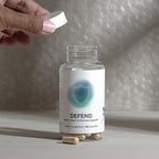 InfiniWell Defend dietary supplement bottle with open cap, visible capsules, and hand holding the bottle lid.