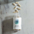 InfiniWell Defend dietary supplement bottle with open top and capsules arranged above on a marble surface.