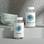 Two InfiniWell Defend dietary supplement bottles with 60 capsules displayed on a white countertop beside stacked bowls.