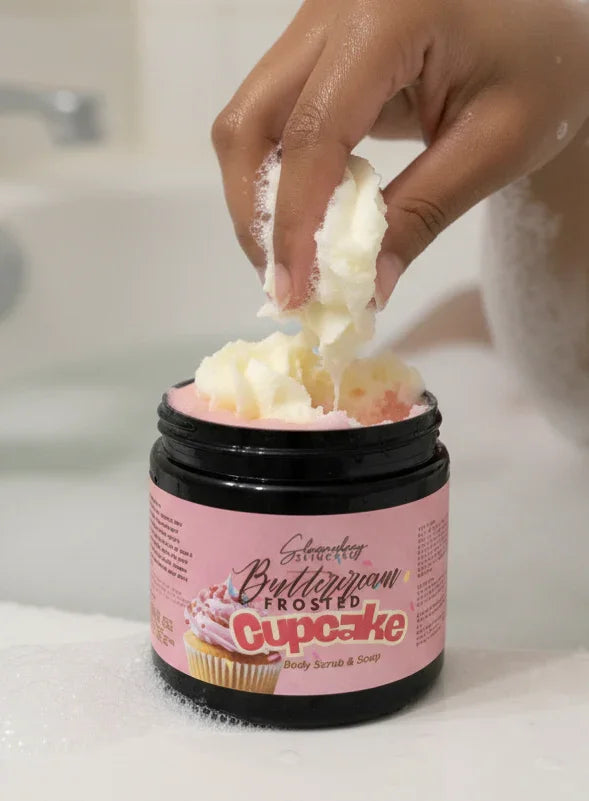 Hand scooping cupcake-scented body scrub from jar in a bathtub for luxury self-care.