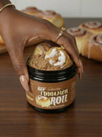 Hand with long light brown nails holding jar of cinnamon roll body scrub and soap on wooden table