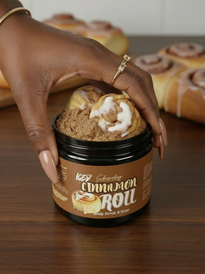 Hand with long light brown nails holding jar of cinnamon roll body scrub and soap on wooden table
