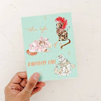 Hand holding a birthday card with decorative cats and text on a light blue background