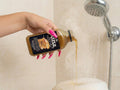 Hand with long hot pink nails pouring maple syrup bubble bath into a tub under shower