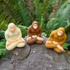 Three collectible Meditating Bigfoot figures are posed on a rock surrounded by ferns, exuding a sense of mindfulness.