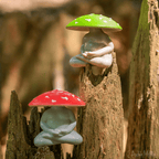 Two Meditating Mushrooms vinyl figurines, one with a red cap and the other with a green cap, rest on tree stumps in a sunlit forest setting. These collectible figures add charm and character to any nature-inspired display.