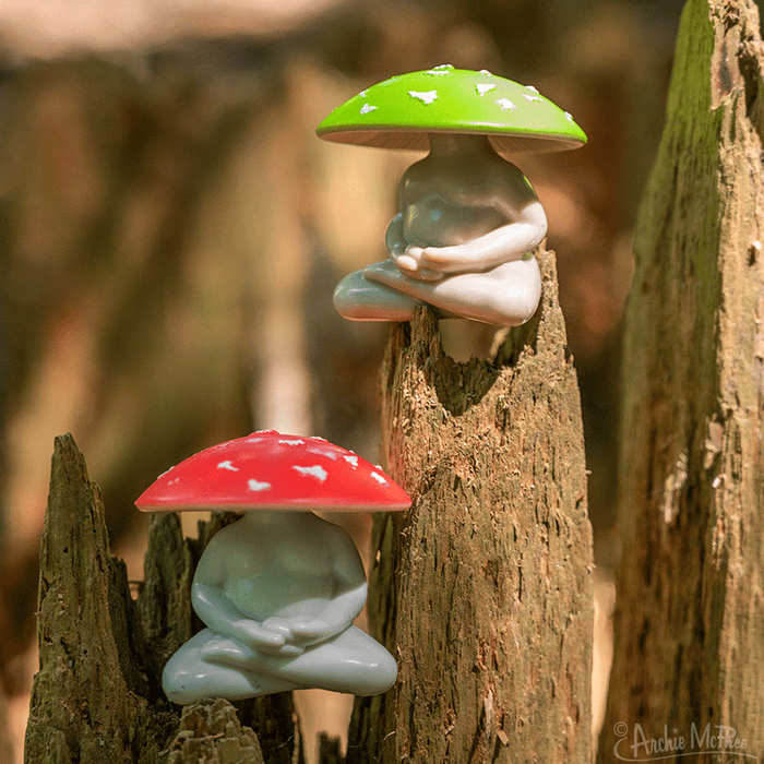 Two Meditating Mushrooms vinyl figurines, one with a red cap and the other with a green cap, rest on tree stumps in a sunlit forest setting. These collectible figures add charm and character to any nature-inspired display.