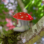The Meditating Mushrooms, a small collectible figure with a mushroom shape featuring a red cap and white spots, rests on a mossy tree branch.