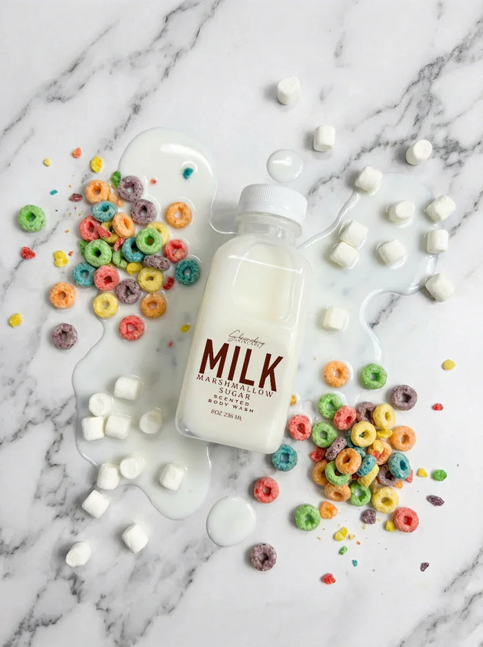 Bottle of marshmallow sugar scented body wash with spilled milk, colorful cereal, and mini marshmallows on marble background