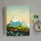 Father's Day card with cartoon mountains on a gray surface with a pen, stamps, and clock.