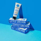 100ml bottle of Little Urchin Natural Clear Zinc Sunscreen SPF 50+ balanced on a stack of its boxes on a blue surface. The sunscreen is a hydrating, vegan formula that provides UVA and UVN broad spectrum protection from the sun while also being 4 hours water-resistant. Packaged in a cream tube with minimal blue and black text on the label.