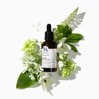 Bottle of hair treatment oil surrounded by green leaves and white flowers on a white background.