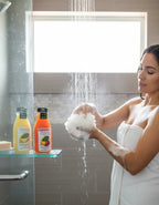 Woman showering with scented body washes, holding a loofah, bottles on glass shelf