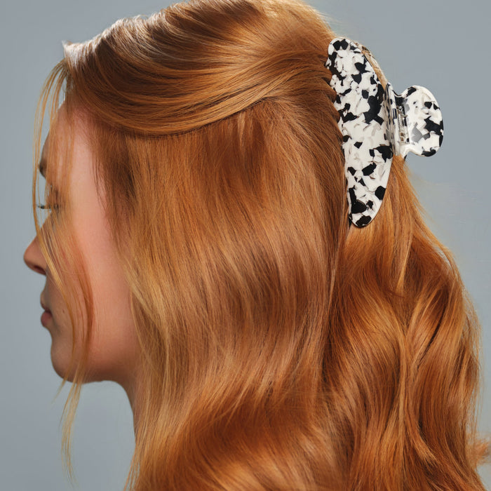 A woman with wavy red hair styled to the side, secured with The Clip by Arey—a large butterfly-shaped, tortoiseshell hair clip that holds all hair types securely—against a plain gray background.