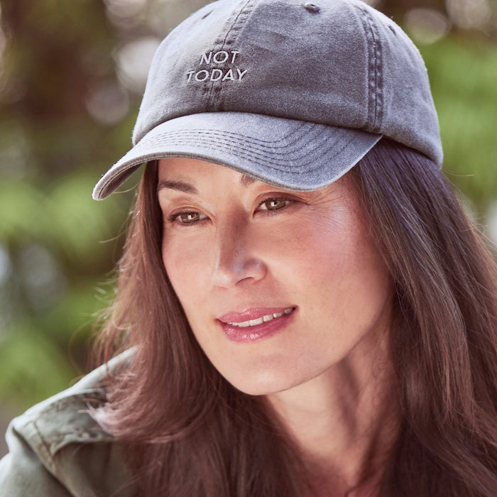 A woman with long brown hair is wearing The Hat by Arey—a gray cotton twill cap embroidered with "NOT TODAY"—as she looks slightly down and smiles softly, framed by a softly blurred green background.